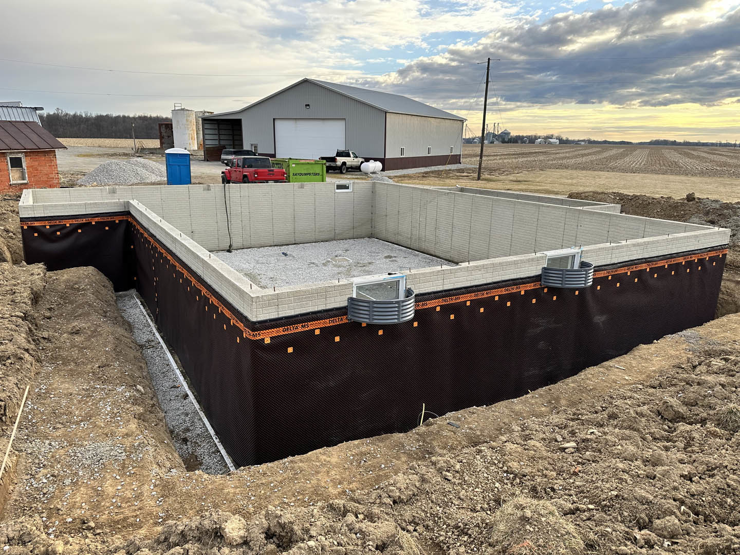Framed custom home under construction with exposed wood beams, brick wall, glass window, red vehicle, blue portable toilet, and truck parked on dirt lot; finished building in