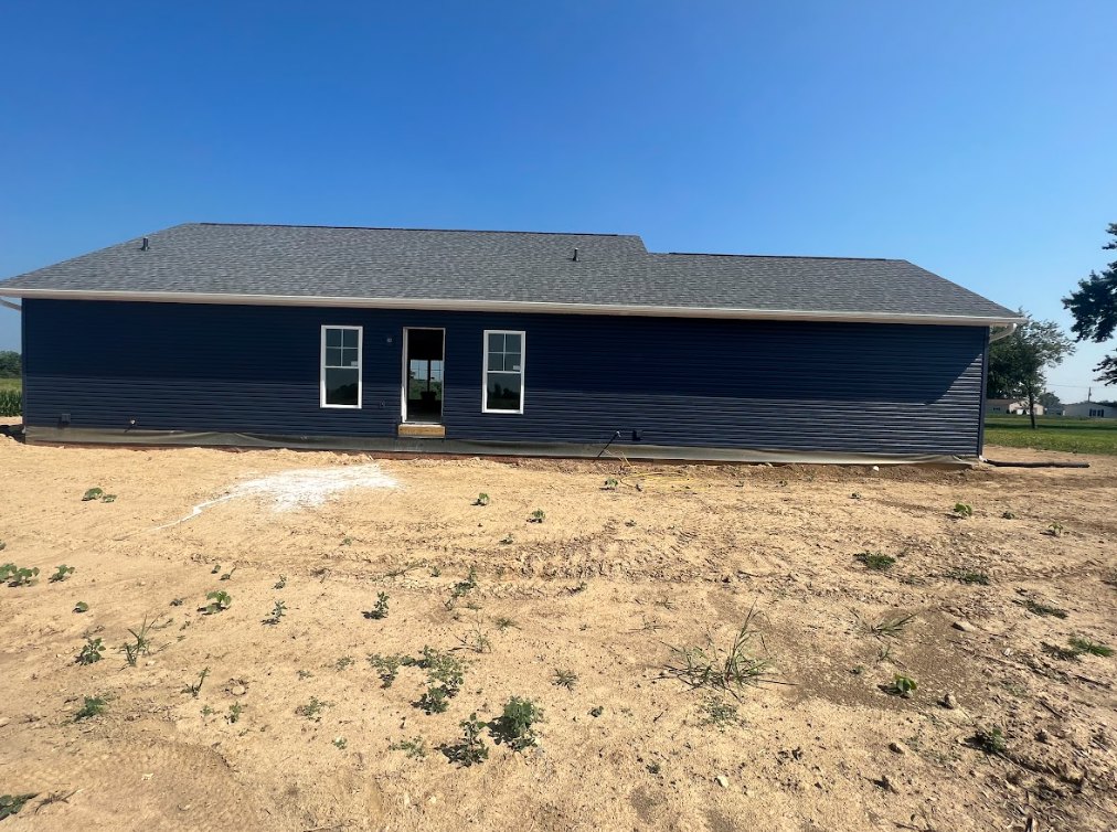 Two-story blue house with white trim and door, unfinished yard with exposed dirt, scattered plants, and clear blue sky overhead