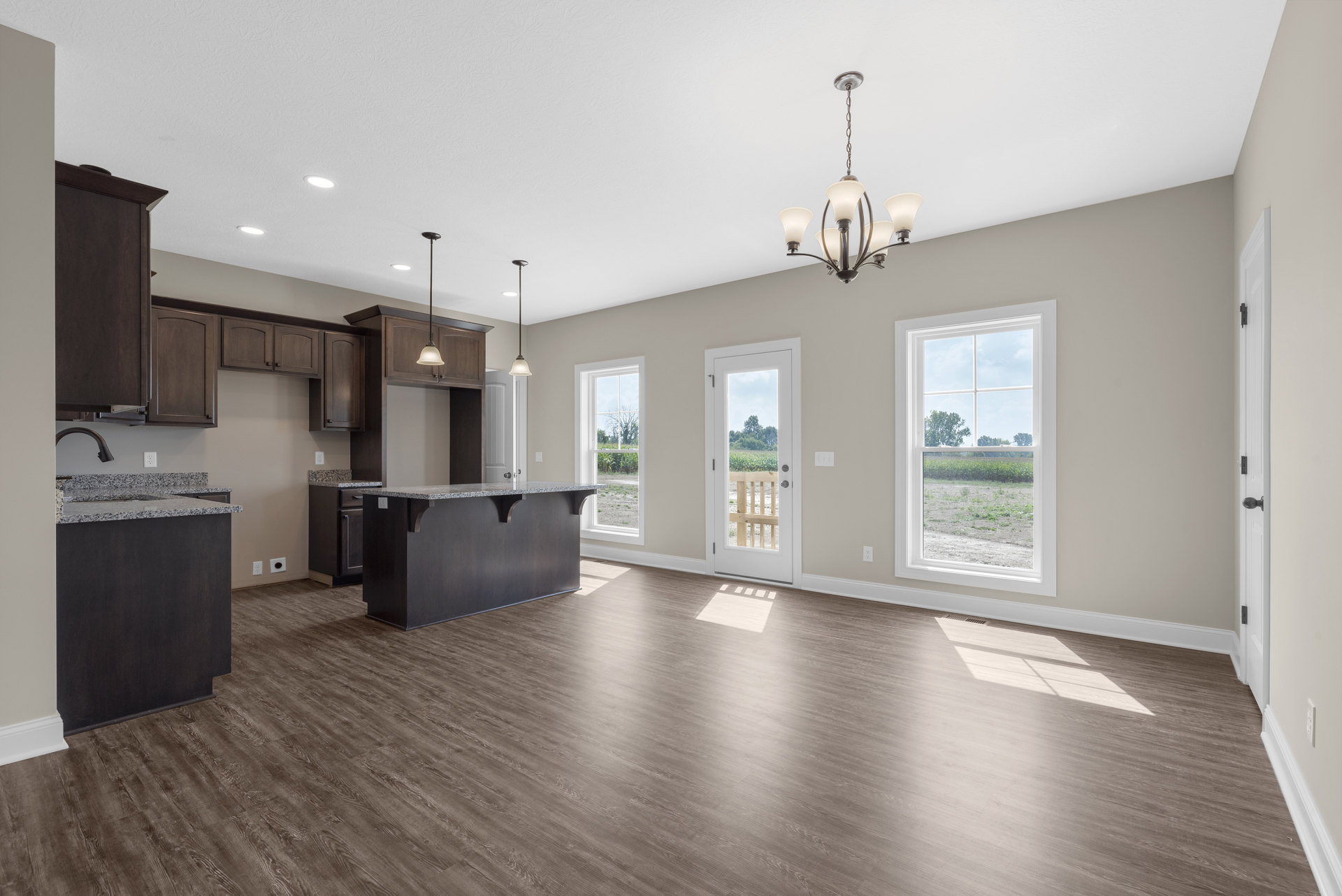 Spacious kitchen with wood flooring, marble-topped island, black and white countertops, cabinetry, chandelier, large window overlooking a field, and door leading to adjacent room