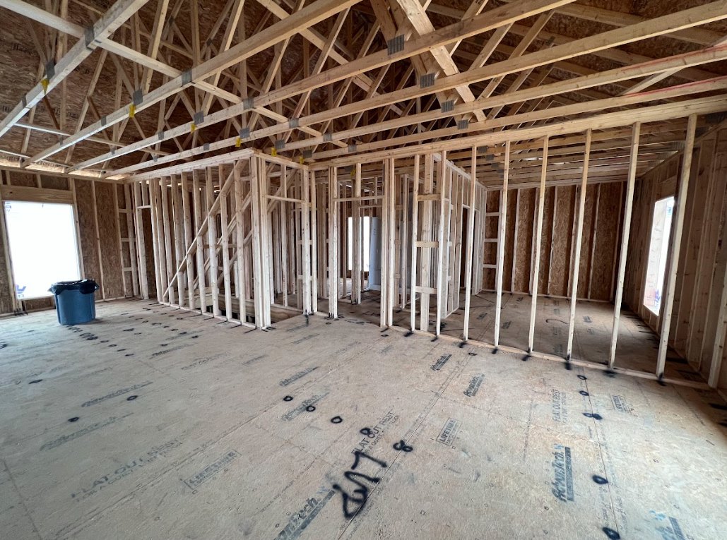 Wood framing and roof structure with exposed beams, blue bucket with black lid on unfinished floor, person standing near window, black-painted wood frame, sunlight streaming