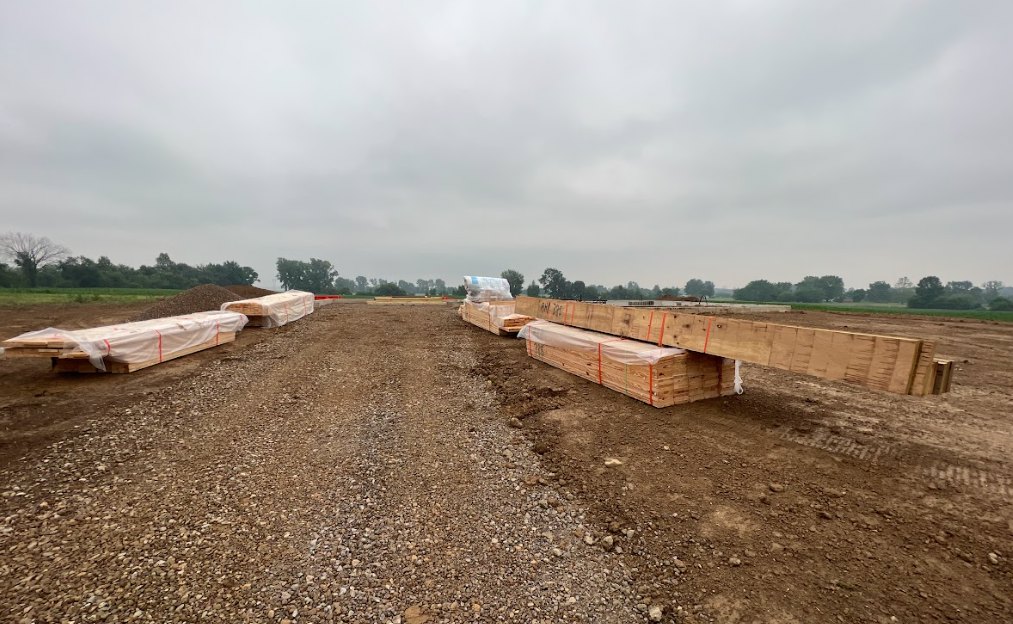 Stack of wooden beams wrapped in plastic sheeting on dirt ground, cloudy sky overhead, grassy field and trees in background