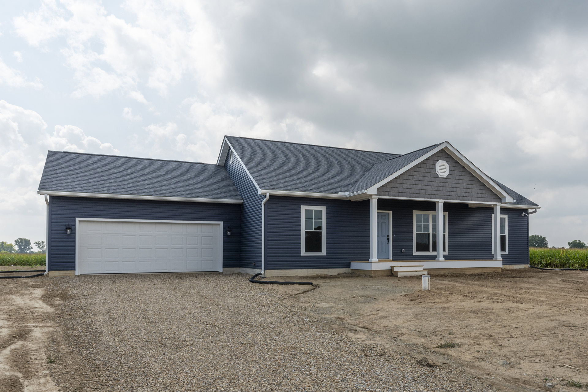 Two-story house with blue roof, white garage door, white-framed windows, paved driveway, and black drainage pipe along the ground.