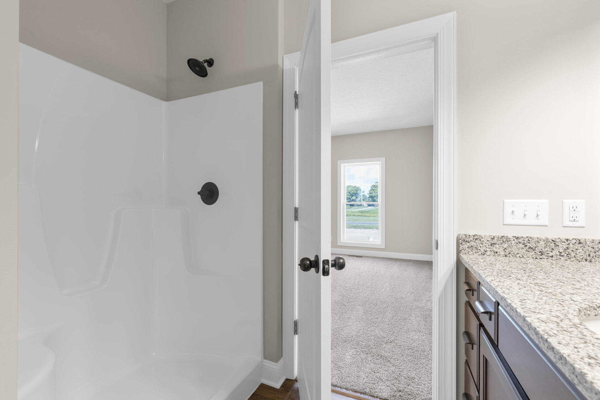 Modern bathroom featuring white tile shower with black spout, white sink, light switch, electrical outlet, and window overlooking trees and grass.