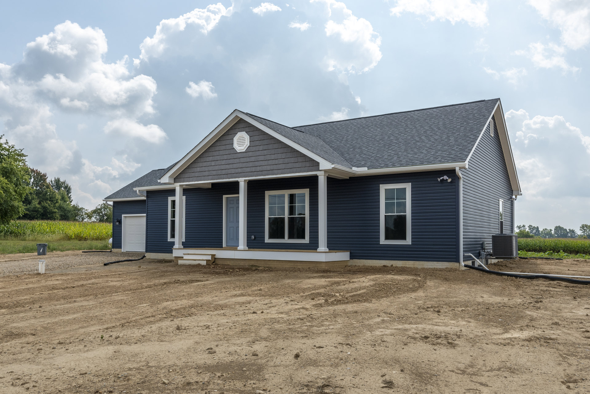Gray house with blue siding, white vent, and windows framed in white trim; dirt ground in front, cloudy sky overhead.