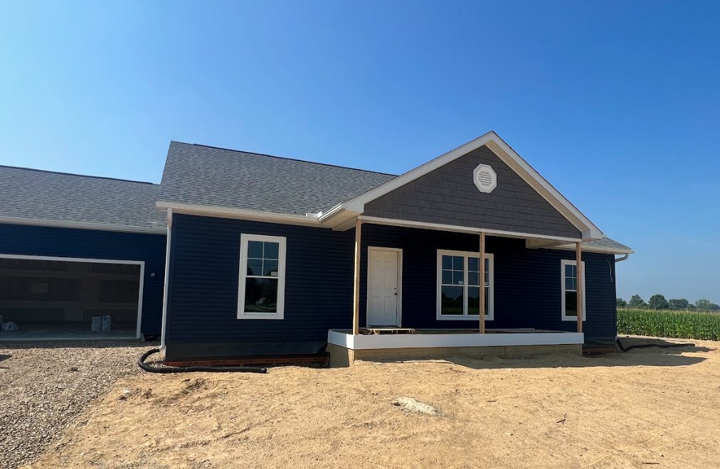 Partially built house with exposed framing, white door, black trim, white-framed windows, covered porch, dirt foreground, blue sky overhead