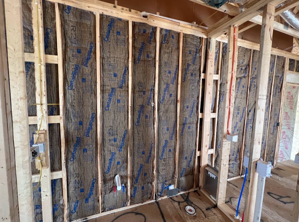 Exposed wood framing with foam insulation panels on walls, white PVC pipe with black text, electrical box with white paper label, construction materials and red writing on paper