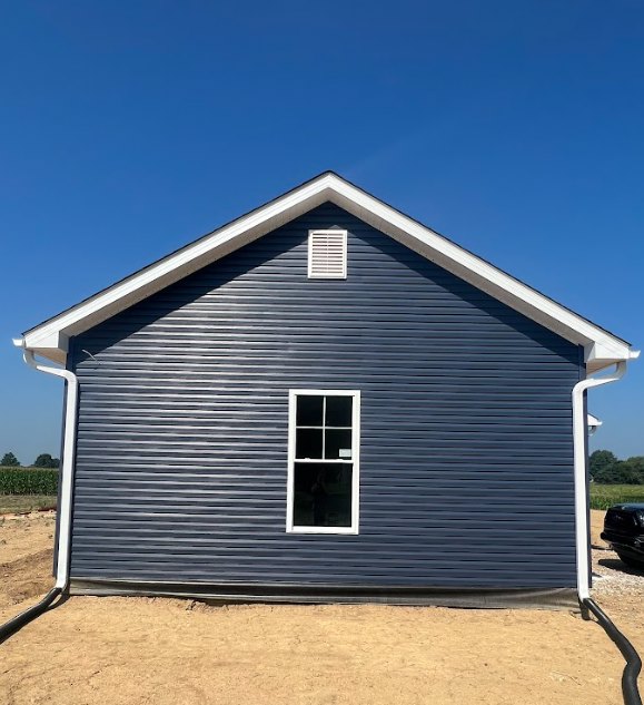 Blue siding house with white roof and white-framed window, roof vent visible, dirt road and field in foreground, clear sky overhead