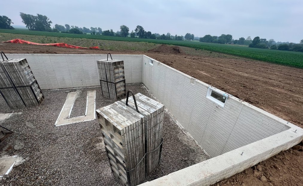 Large concrete foundation surrounded by soil and grass, wooden crates stacked and secured with a chain, pallets on a dirt road, red construction equipment on the ground, trees and