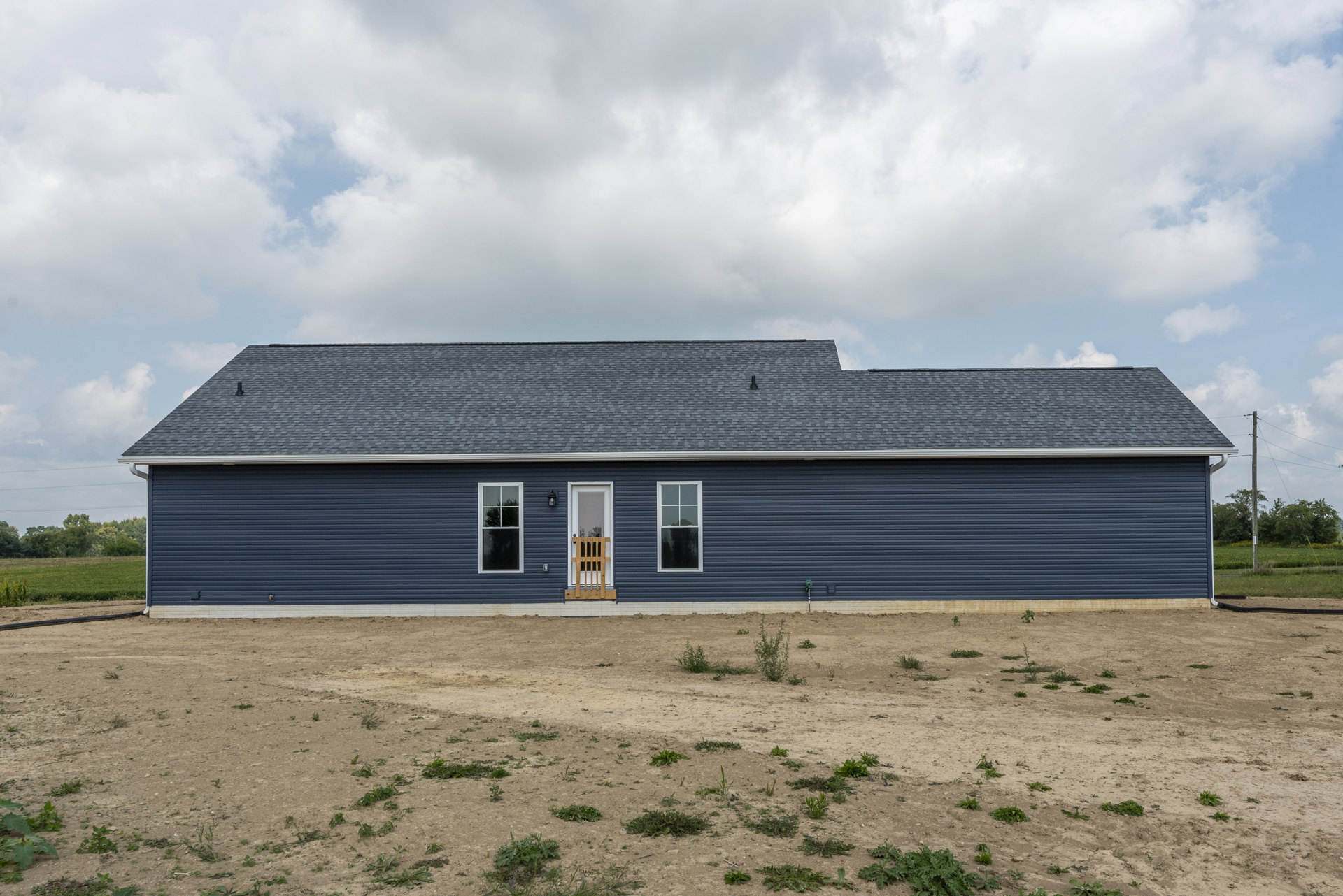 Blue cottage with white-framed windows reflecting trees, wooden front door, gray roof, and dirt field with scattered green plants under a cloudy sky
