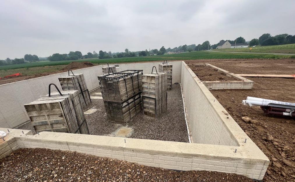 Concrete foundation with several piles of concrete, white pipes protruding, wooden crates stacked nearby, brick wall visible in foreground, grassy outdoor setting with trees and