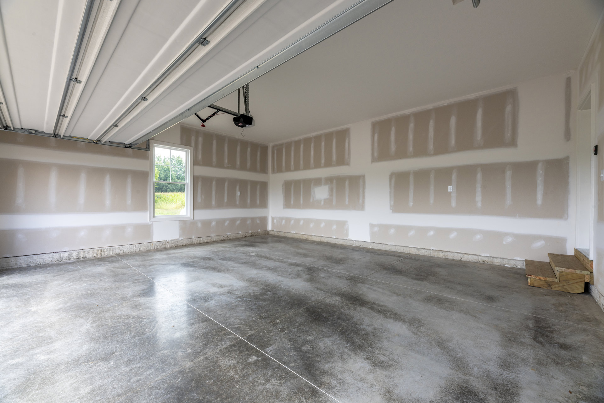White-walled room with grey concrete floor featuring white lines, window overlooking trees, and wooden staircase with a few steps.