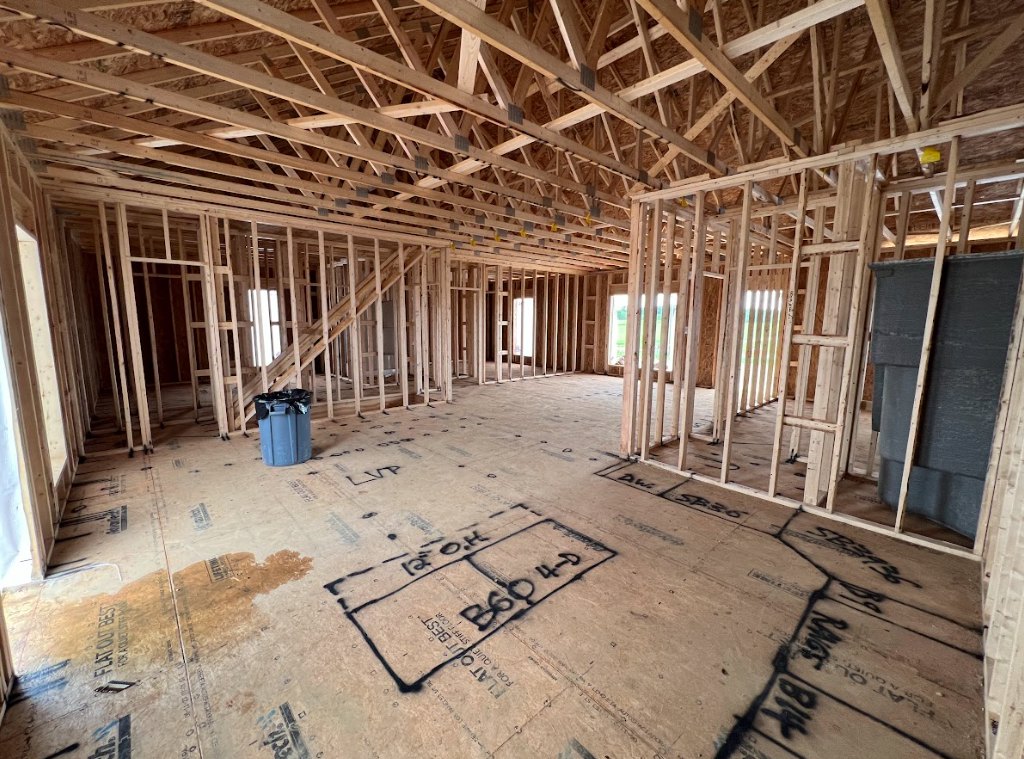 Exposed wood framing and roof trusses inside a house under construction, cardboard with black writing, grey trash can with black bags, black barrel, wooden ladder, and floor with
