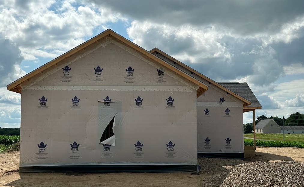Partially built house with white weatherproof sheeting featuring blue logos, exposed framing, large opening in the wall, pile of rocks on dirt ground, cloudy sky in background