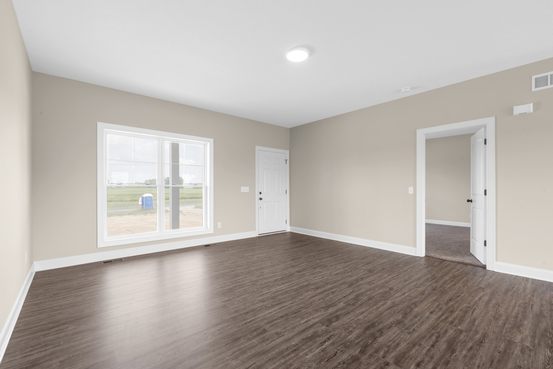 Wood flooring in a room with white walls, a white door with black knobs, ceiling light fixture, wall vent, and a window overlooking a field; blue toilet visible near the window.