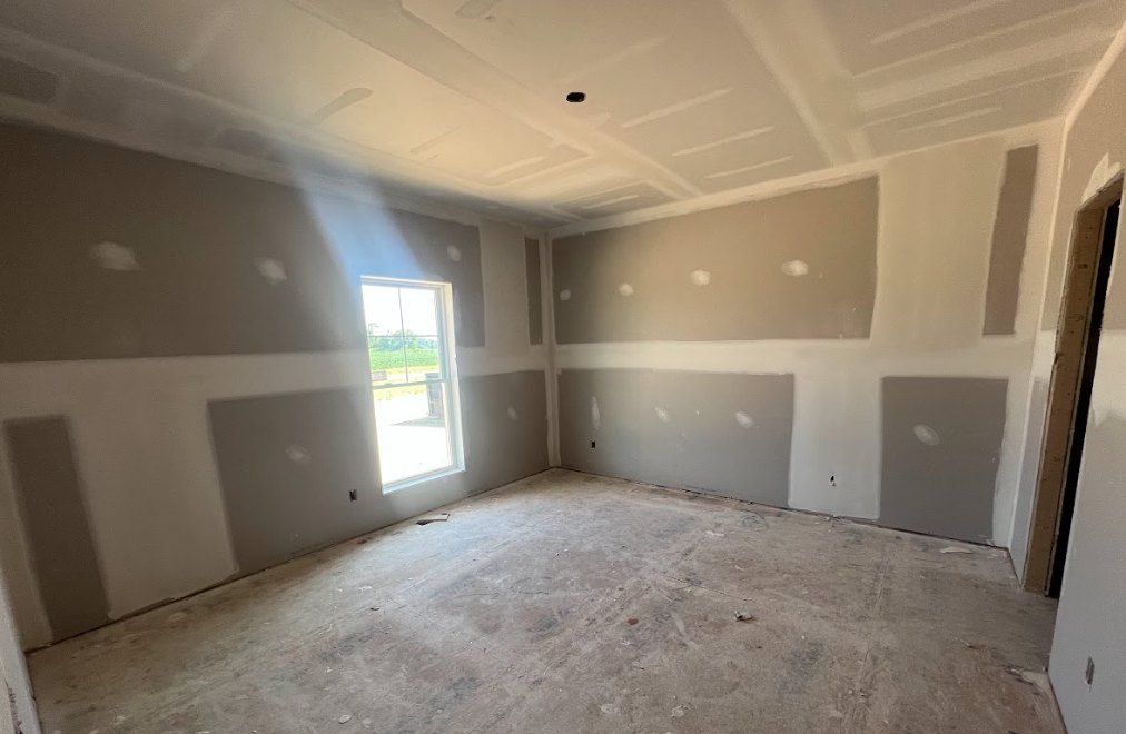 White-walled room with large window overlooking a grassy field, concrete floor, ceiling opening, and suspended white fixture
