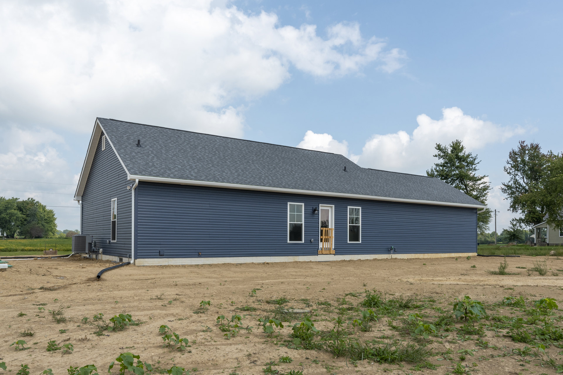 Blue house with white-framed windows and door, wooden railing in window, dirt field with sparse plants, tree nearby, cloudy sky overhead.