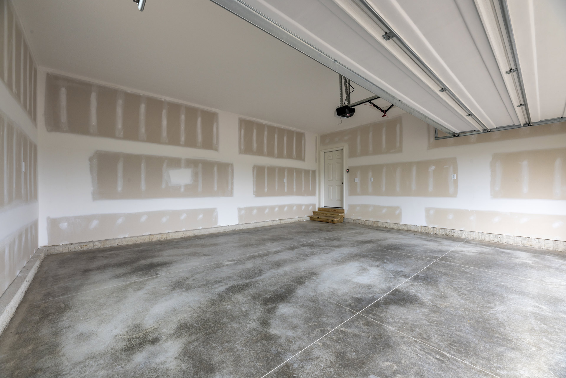 Garage interior with concrete floor, wooden step, white walls, open white door, grey tile flooring with white lines, wooden stairs in front of window, and closed garage door