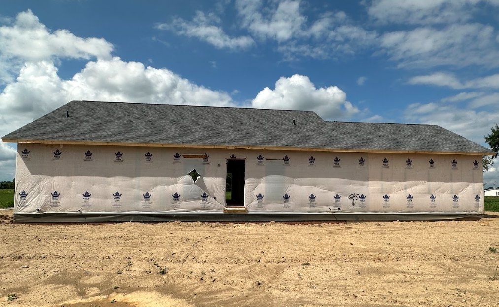 Partially built house with exposed plywood walls, large opening in the exterior, black doorway, white construction sheet, dirt yard, green lawn, and cloudy sky above
