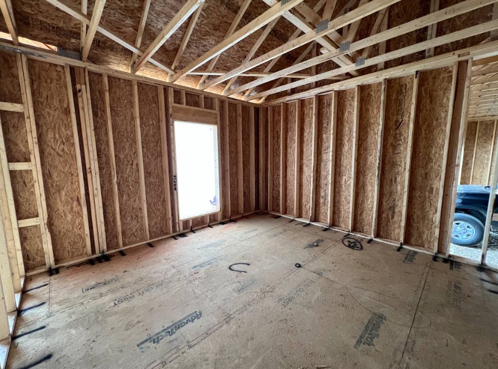 Concrete floor with exposed wood ceiling beams, large window framed in white, unfinished walls, natural light illuminating interior construction materials