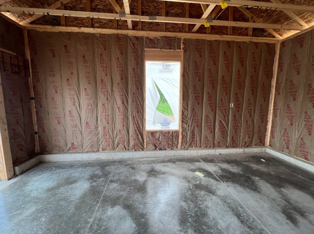Concrete floor and unfinished walls with brown paper covering, large window, exposed wooden ceiling beam.