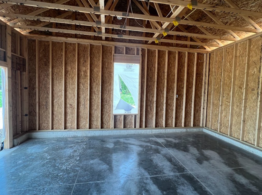 Room with exposed wood ceiling beams, large window with sheer white curtain, smooth concrete floor, and unfinished wall surfaces