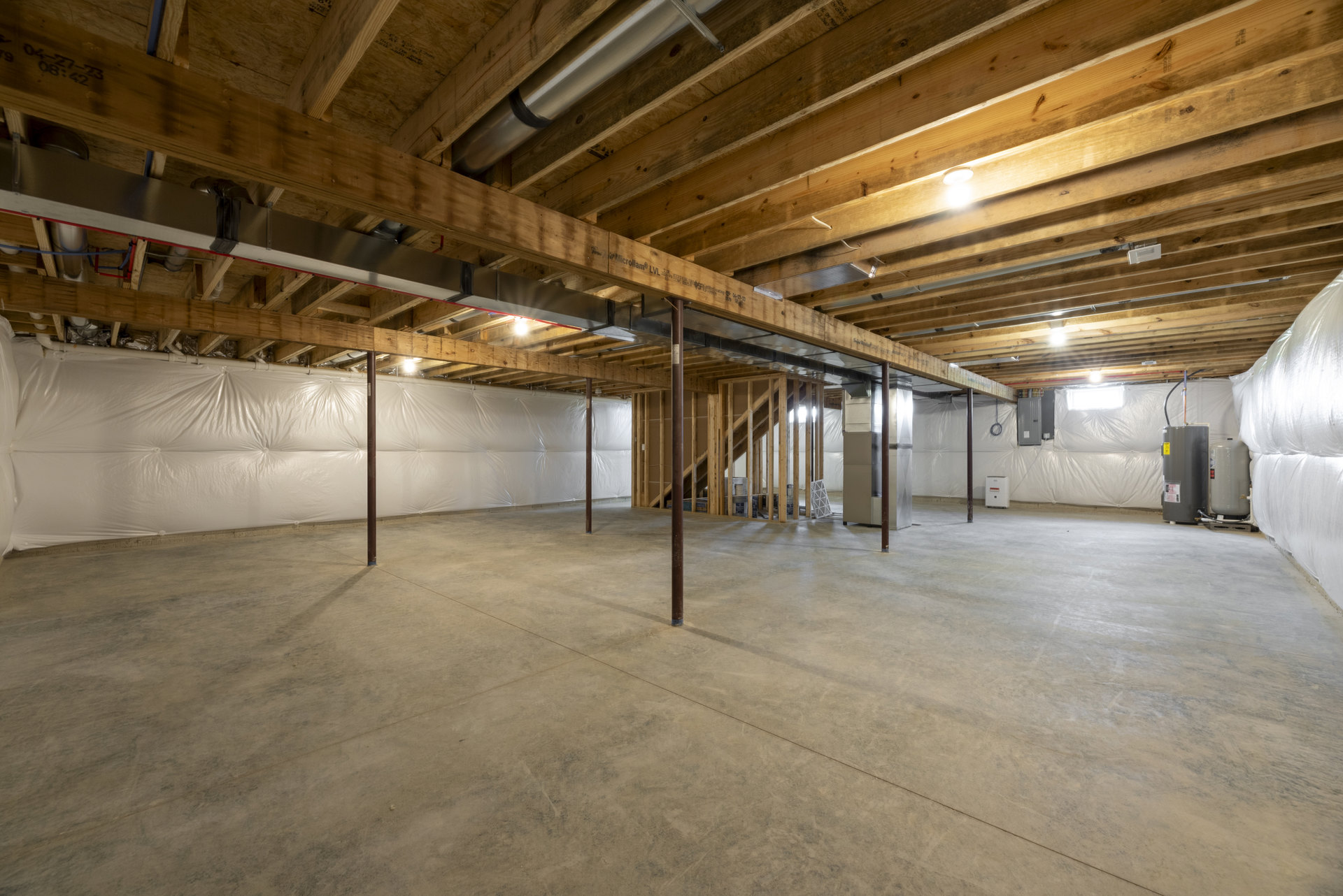 Exposed wooden ceiling beams, concrete floor with central metal pole, industrial light fixture, white plastic wall covering, unfinished basement interior