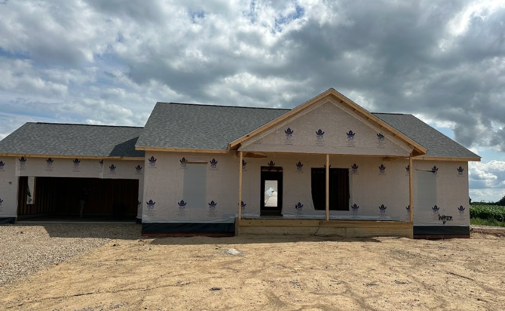 Partially built house with exposed framing, unfinished roof, and construction materials scattered on the ground beneath a cloudy sky