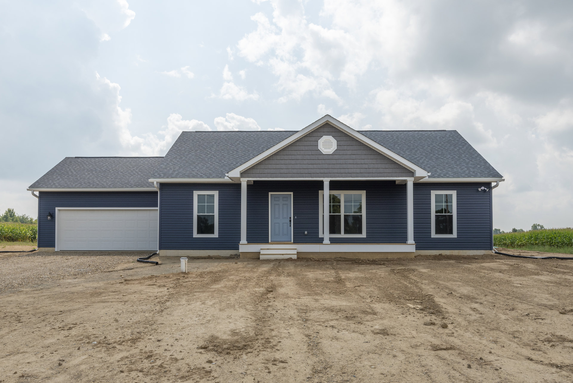 Blue house with white trim, blue front door, white porch, dirt driveway, gray exterior walls, white-framed windows, cloudy sky overhead