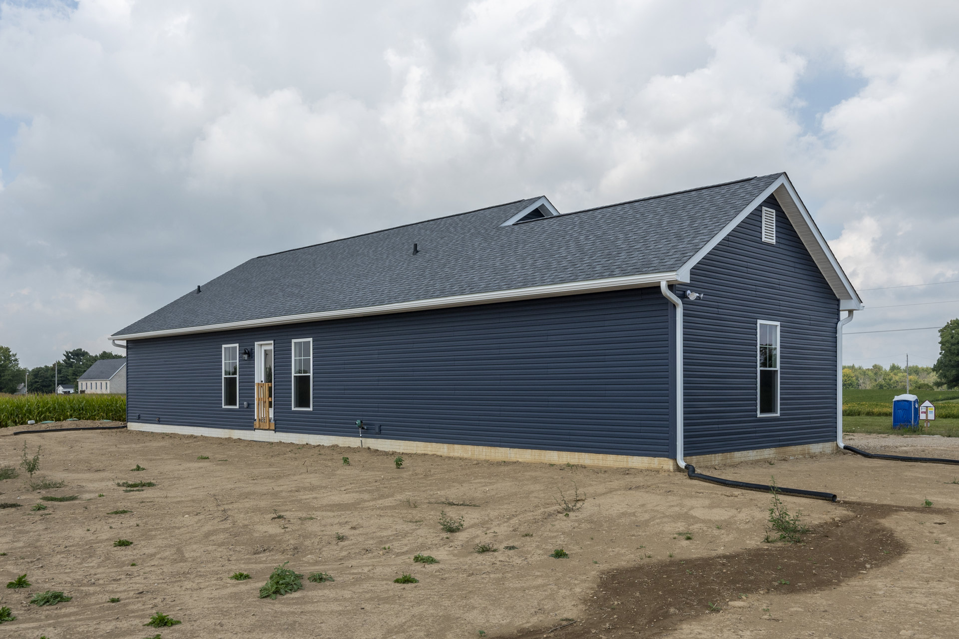White siding house with blue metal roof, surrounded by dirt field with small plants, under partly cloudy sky.