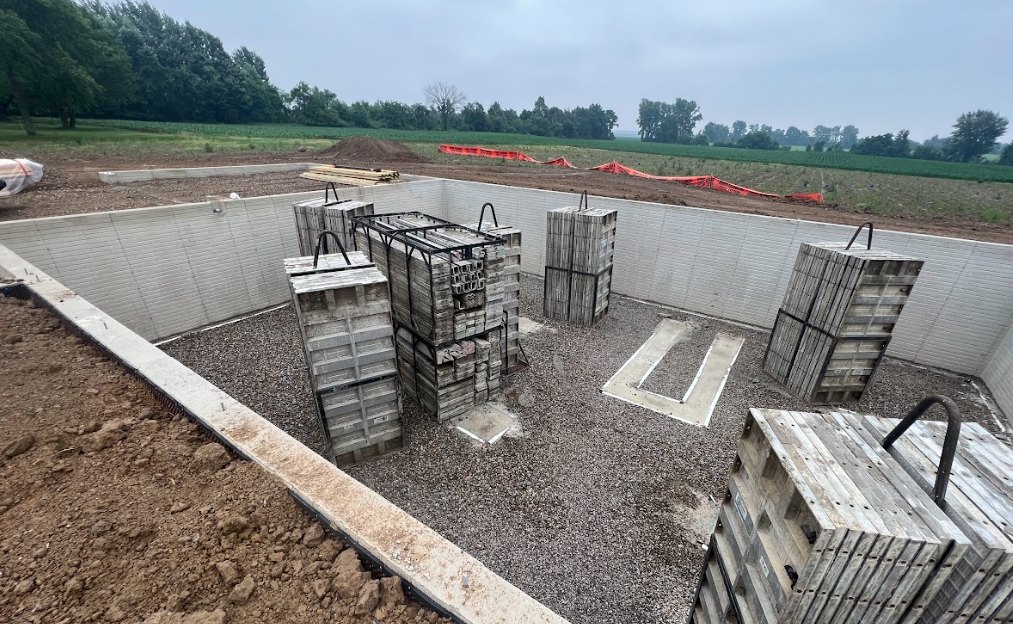 Construction site with stacked wooden pallets, concrete slabs, red tarp on soil, partially built foundation, trees and fence in background