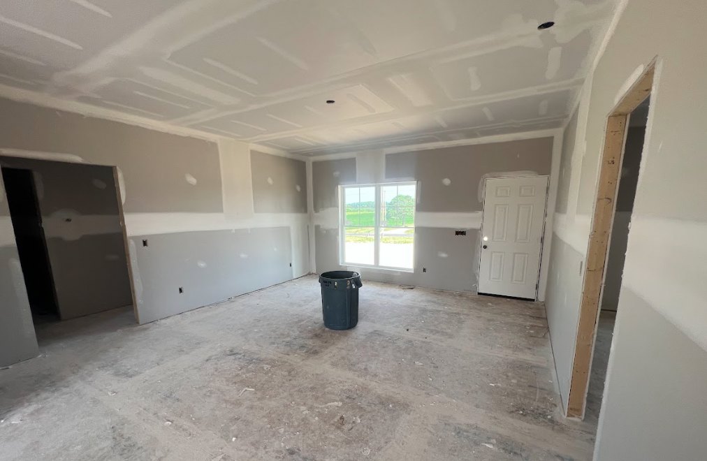 Black trash can on unfinished concrete floor, white door with black handle, window overlooking grassy field, wall and ceiling with visible holes, plaster surfaces, modern interior