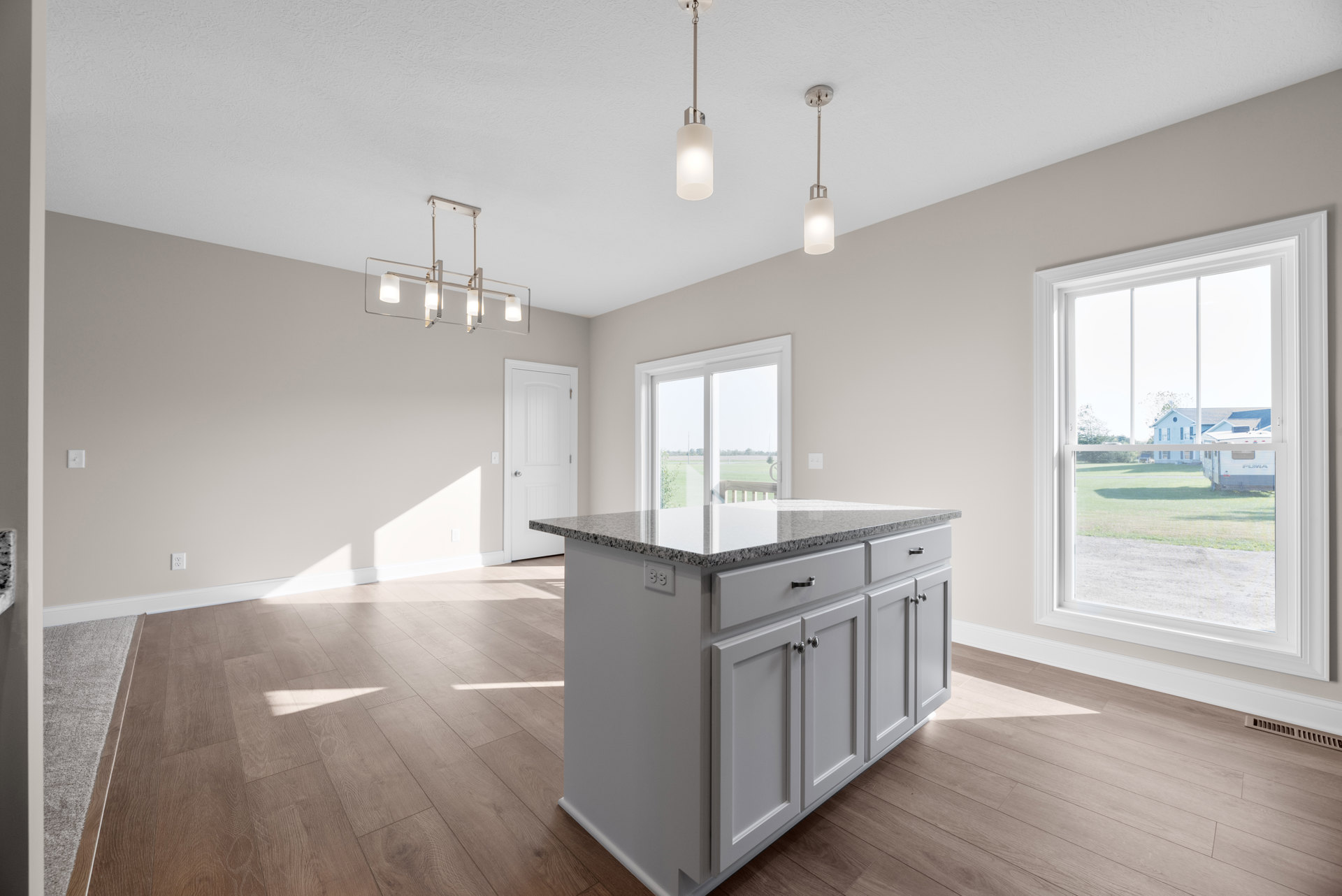 Granite-topped kitchen island with white cabinetry, pendant light fixture above, stainless steel sink, hardwood flooring, and large window overlooking lawn and neighboring house
