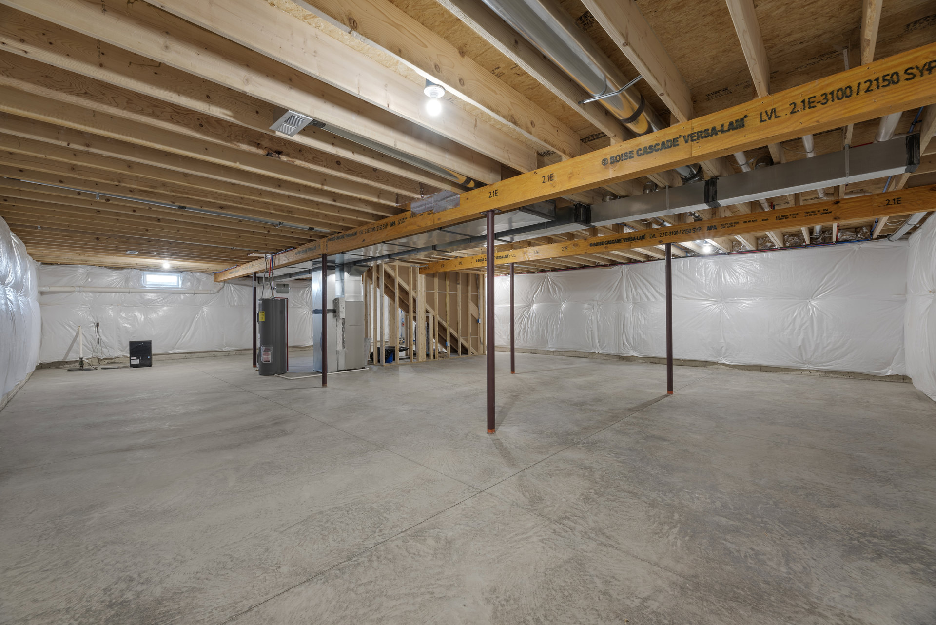Open room with exposed wooden ceiling beams and a central metal beam, concrete floor with a metal support pole, white bed in foreground, single ceiling light bulb, large silver