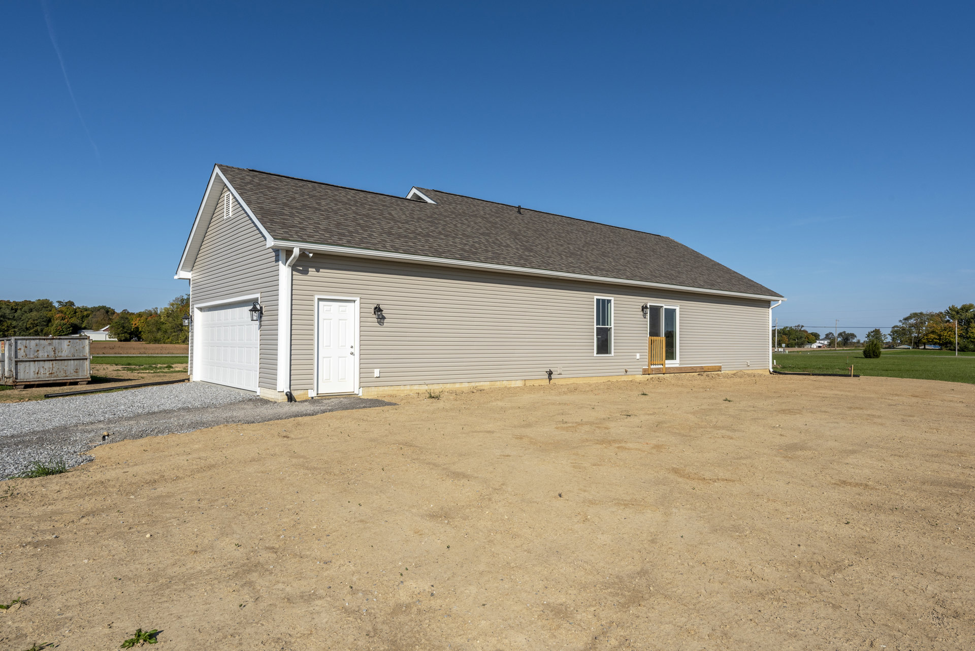 Single-story home with light siding, white front door featuring silver handles, dirt driveway and yard, metal container near entrance, gable roof, sparse landscaping, and open sky.