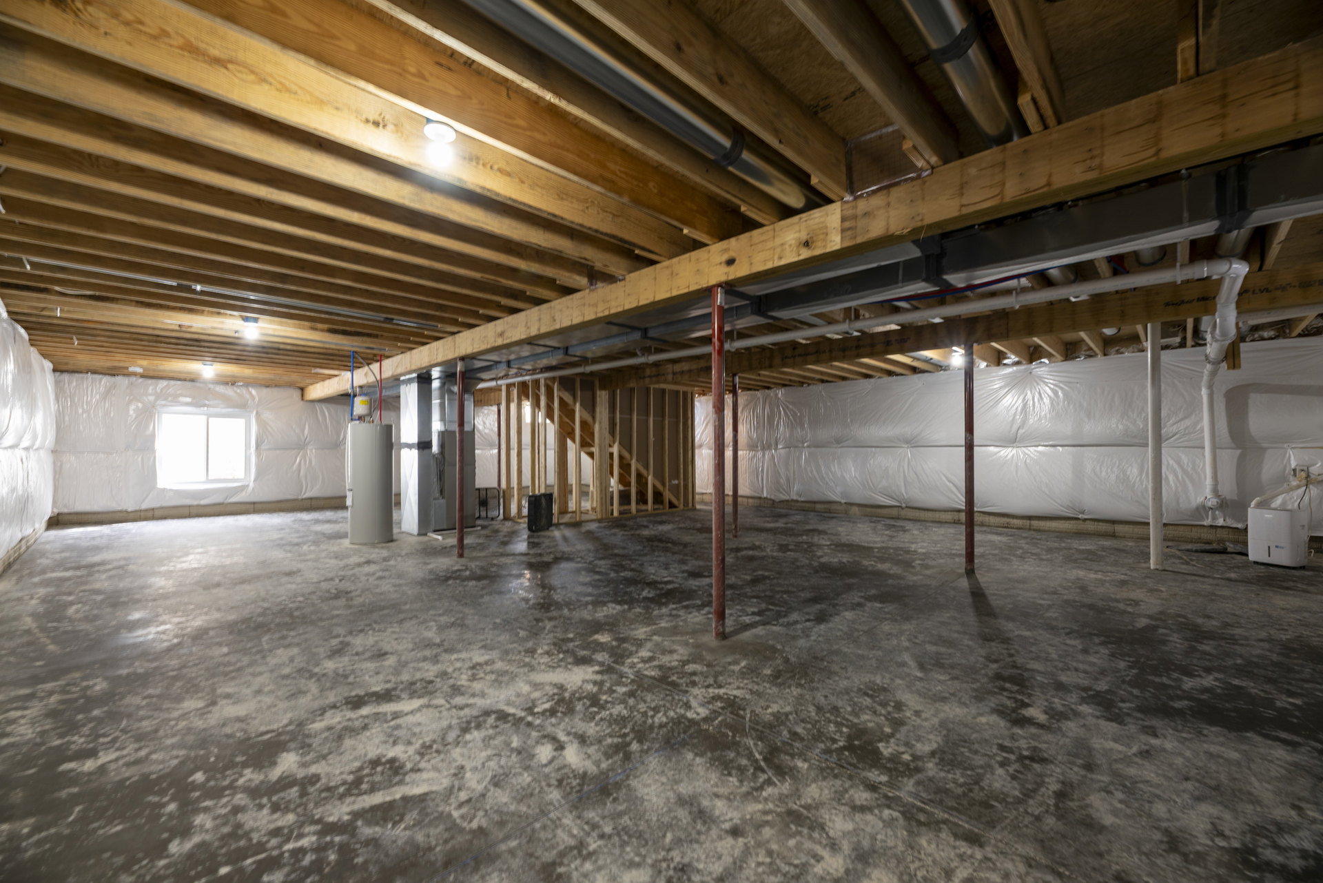 Basement room with exposed wooden ceiling beams, white walls, concrete floor, red and white support poles, metal pipe, window covered in plastic, and a white rectangular object