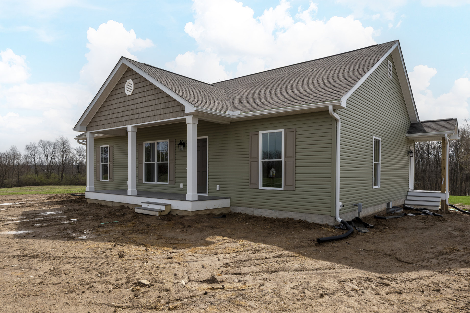 Two-story house under construction with white framed windows, unfinished porch, exposed siding, dirt yard, and clear blue sky