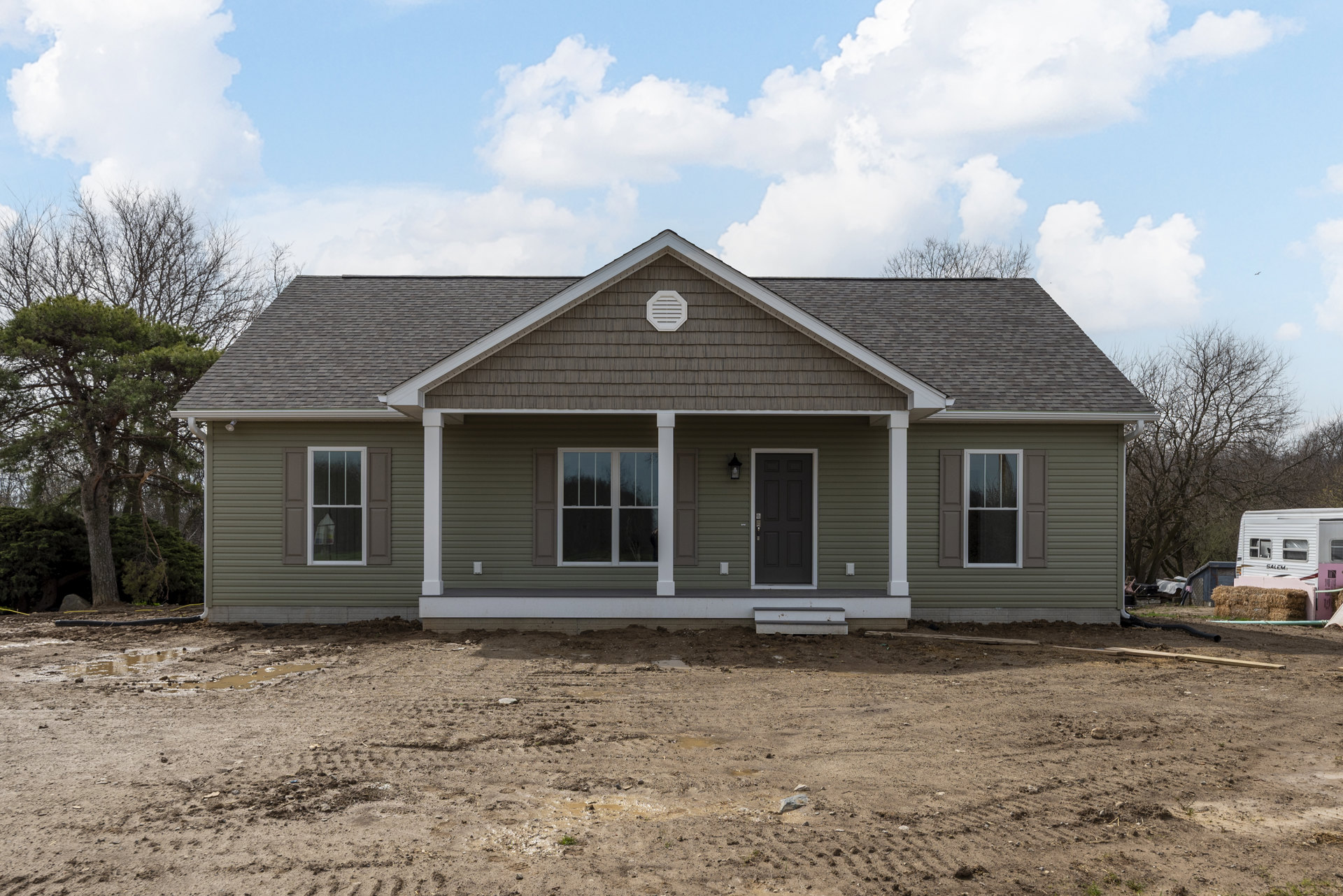 Brown exterior walls with white vents, white-framed windows, and a black door with silver handle; covered porch, dirt yard, and surrounding trees under a cloudy sky.