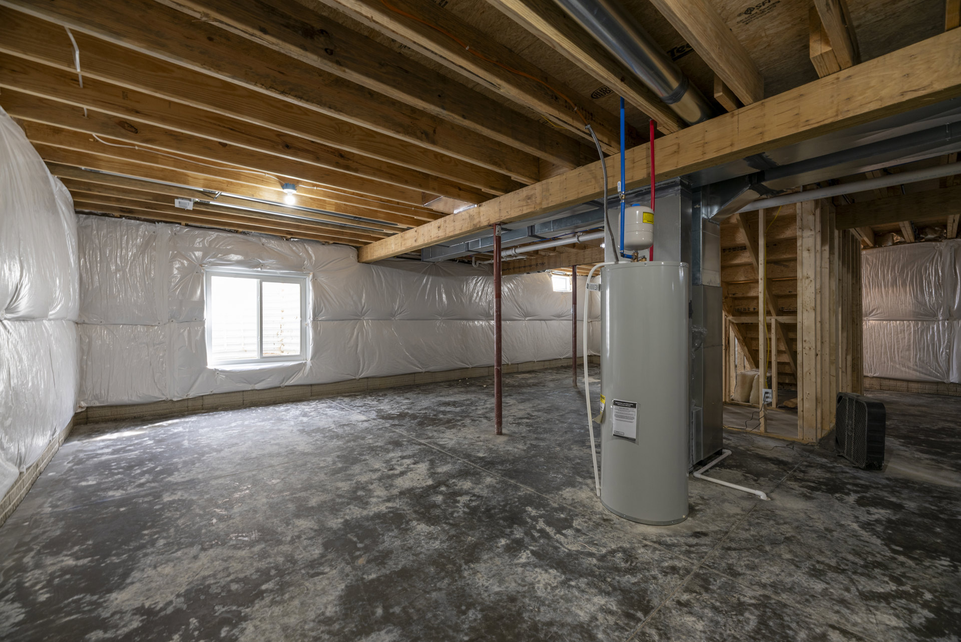 Basement utility room with exposed pipes, white cylindrical water heater, plastic-covered window, unfinished plaster walls, and visible ceiling beams.