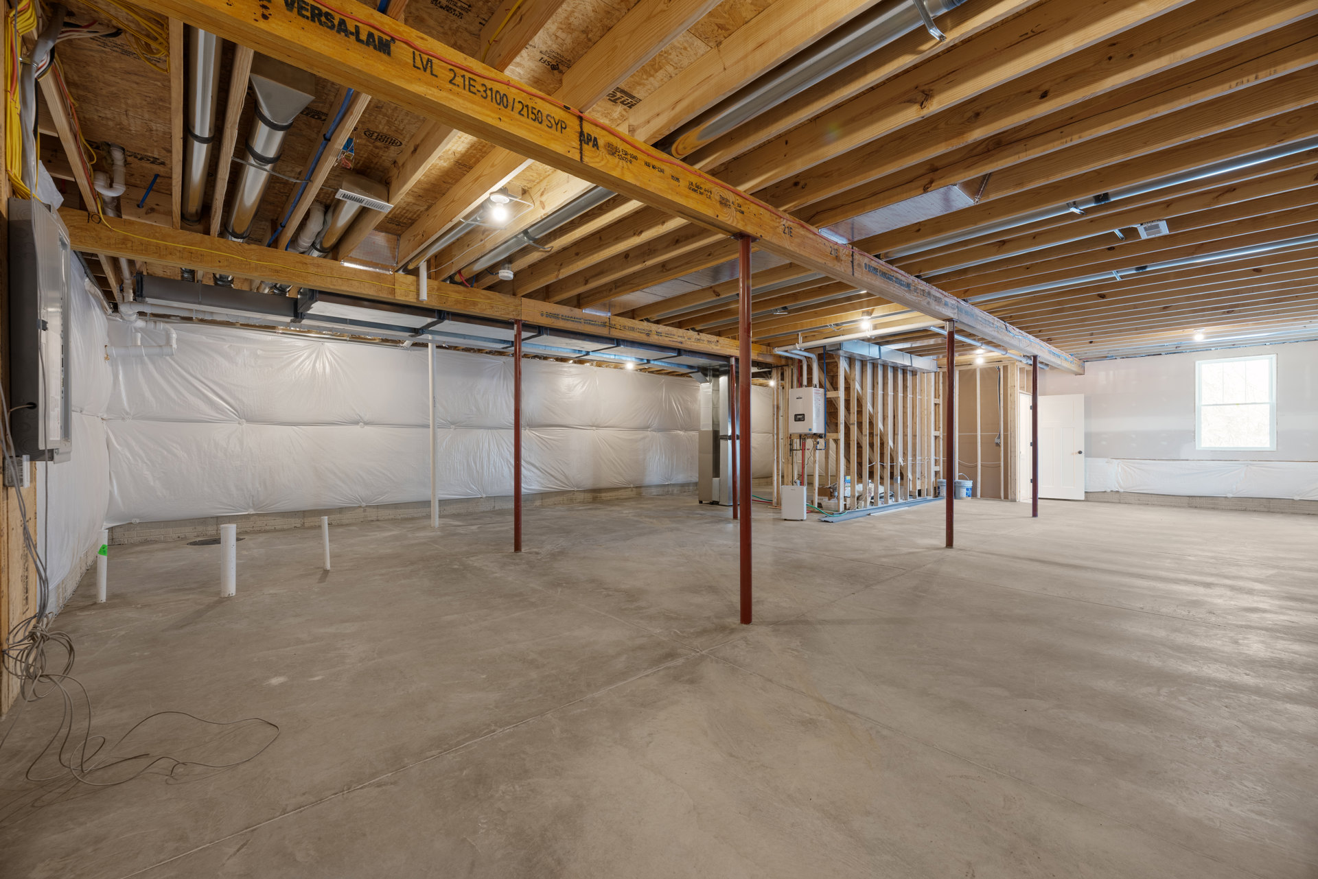 Wooden ceiling with exposed beam, white-framed window, unfinished concrete floor with visible pipes, bare walls in a basement room.