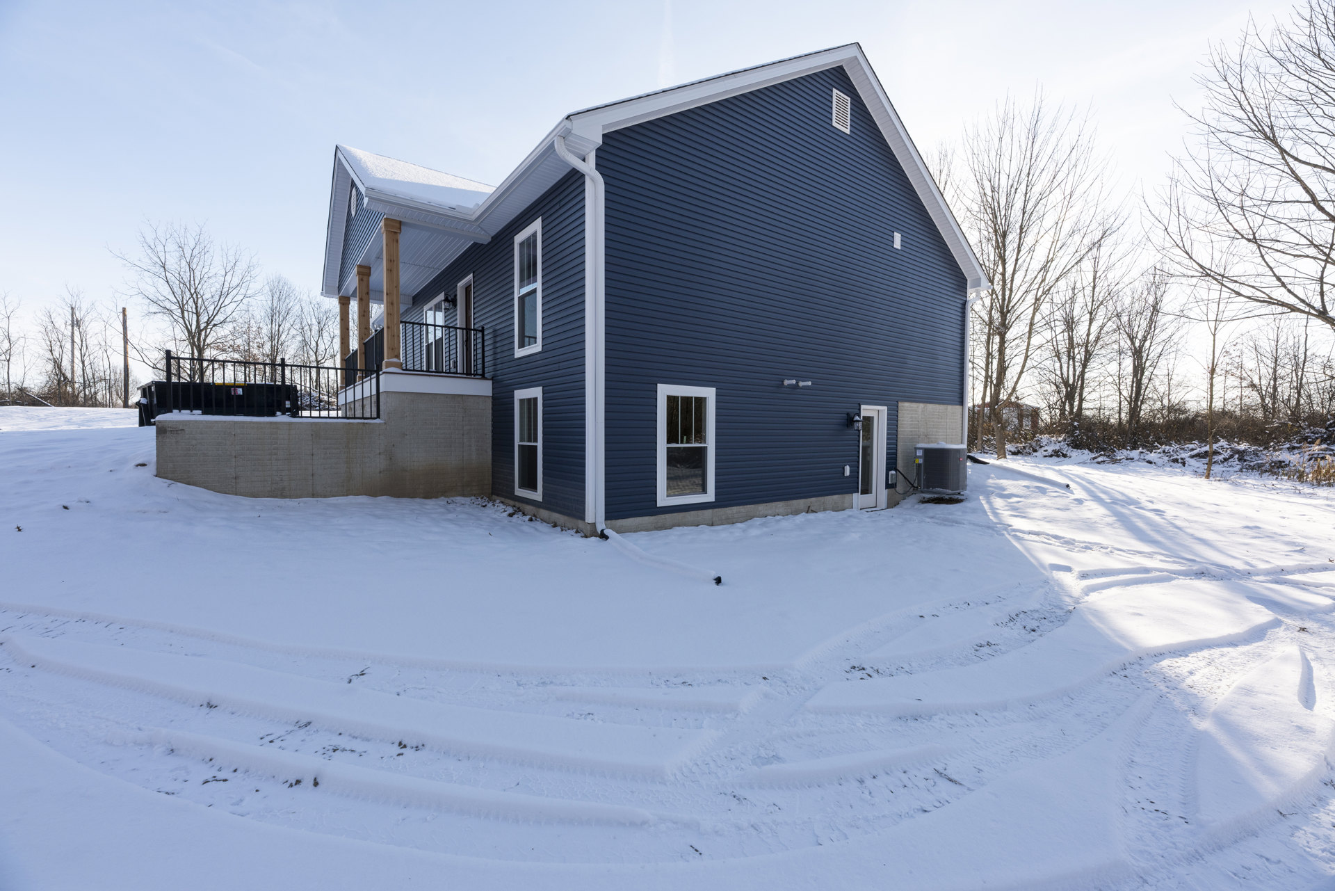 White snow covers the ground around a house with white-framed windows, barred window, and metal fence; large air conditioner sits beside the home's side exterior.