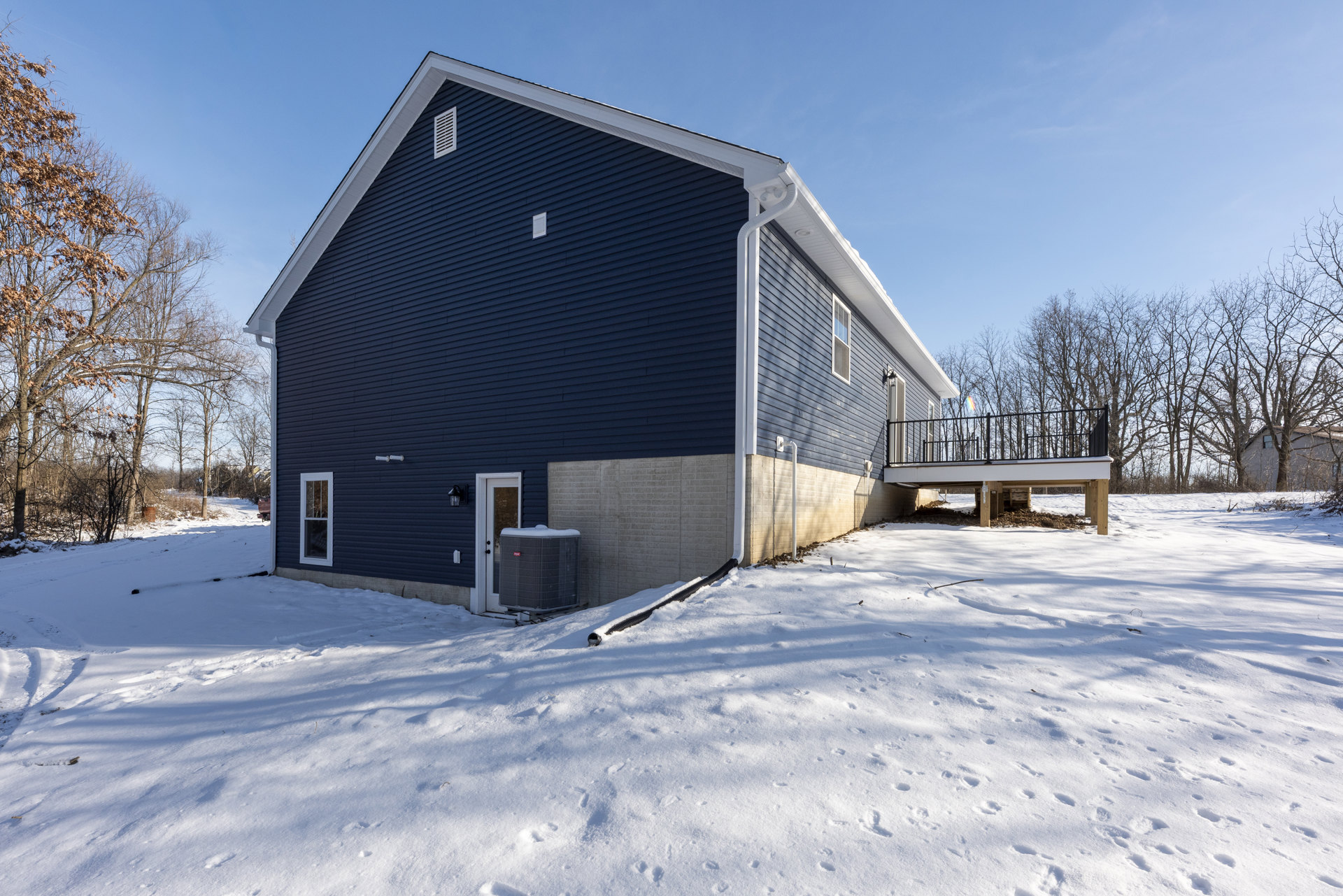 Blue house with white-trimmed deck, snow covering ground and deck, brown-leaved tree nearby, large air conditioner unit beside door