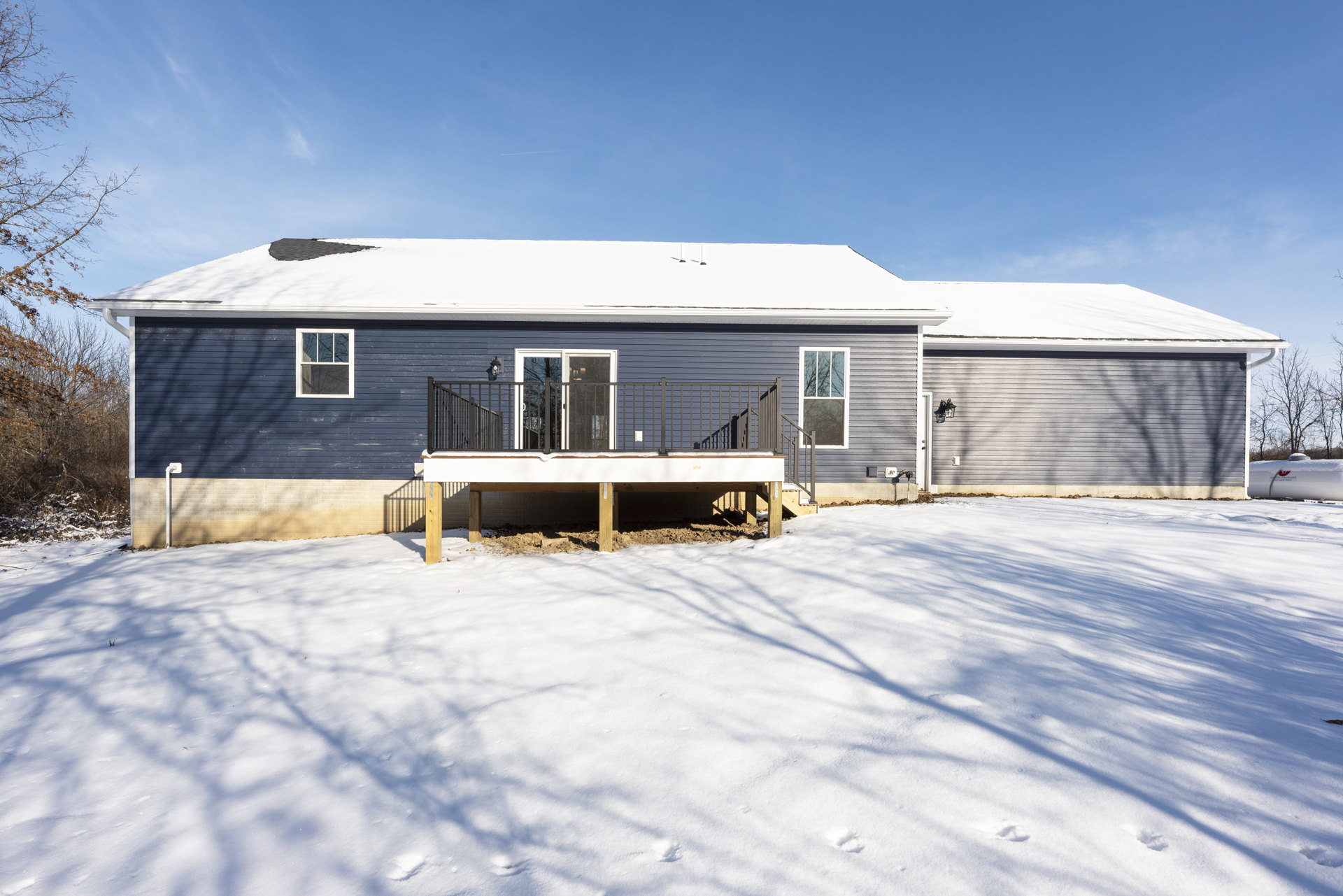 Two-story house with white-framed windows, wooden porch posts, deck with railing, surrounded by snow-covered ground and leafless trees under a cloudy winter sky