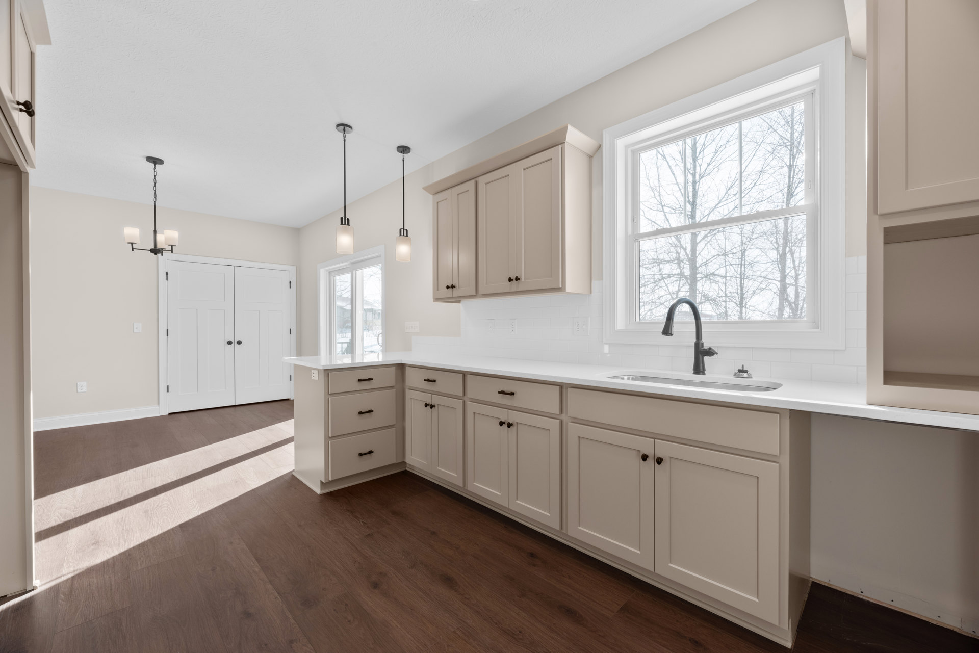 White kitchen cabinets, black undermount sink with matching faucet, white double doors with black knobs, window overlooking trees, light-colored countertops and walls