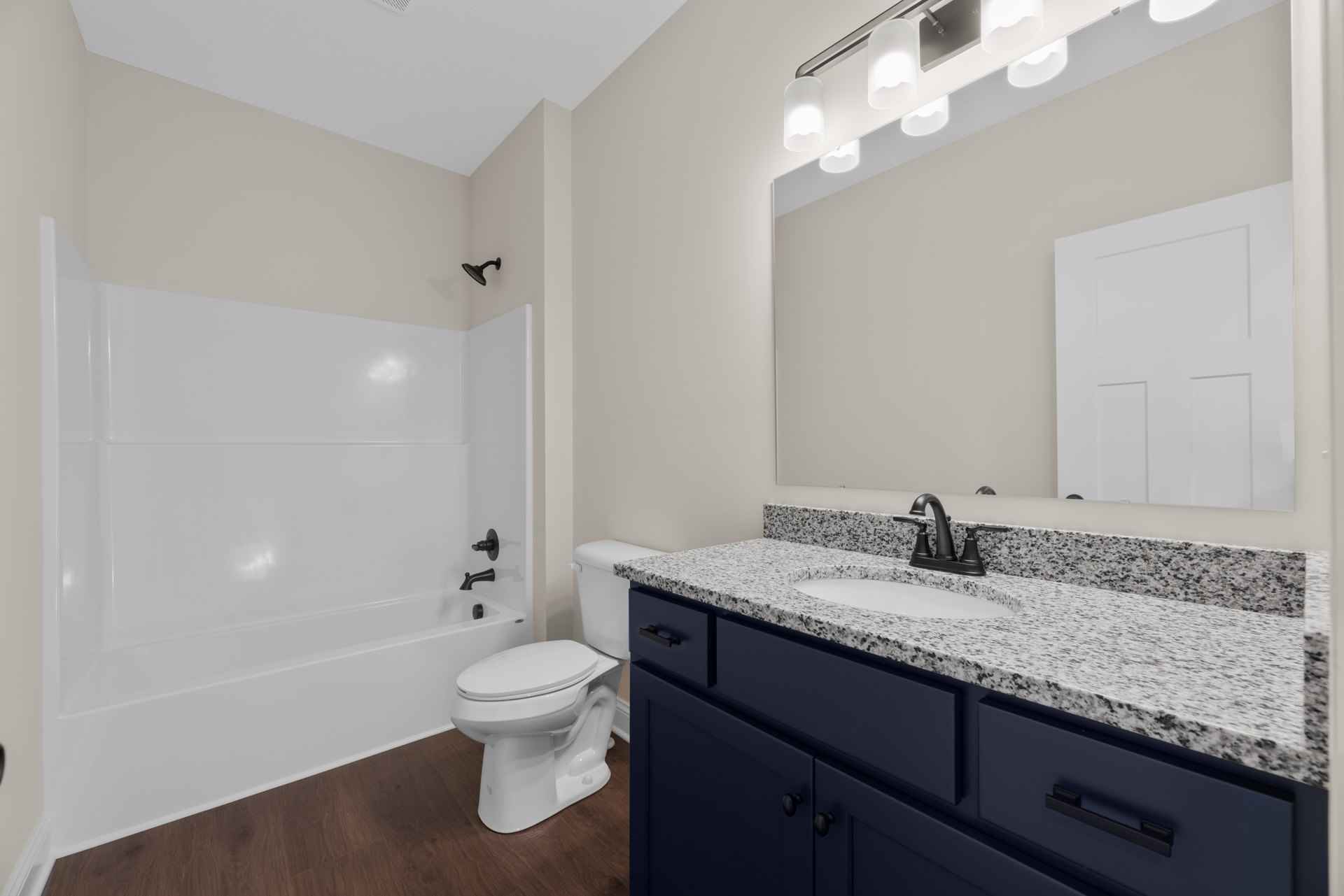Bathroom with white tile floor, white bathtub and toilet, rectangular mirror above a sink with cabinetry, white door with square panel, modern light fixture illuminated above the