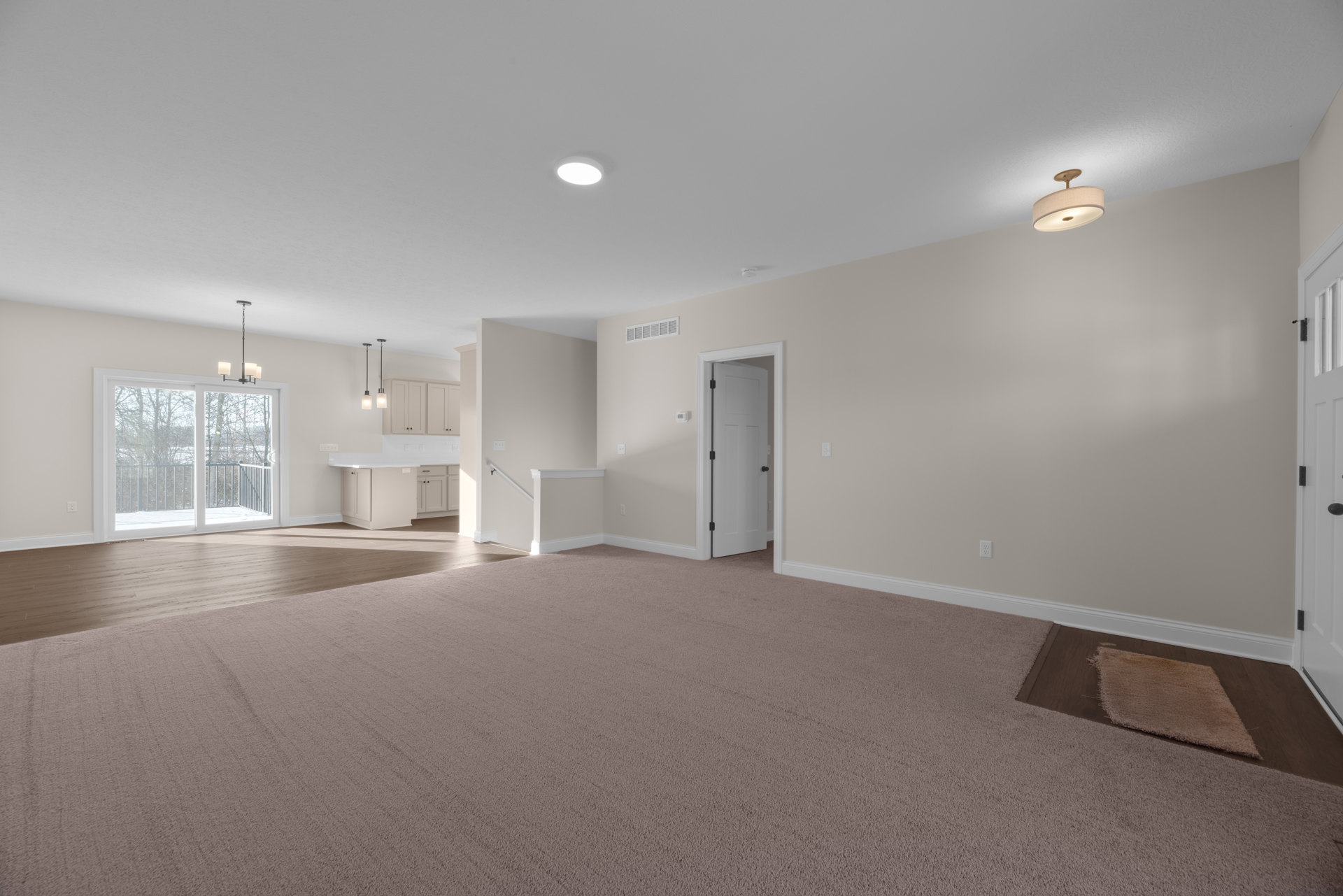 Spacious carpeted room with white walls, beige flooring, a white door, window with railing overlooking trees, and ceiling light fixture with shade