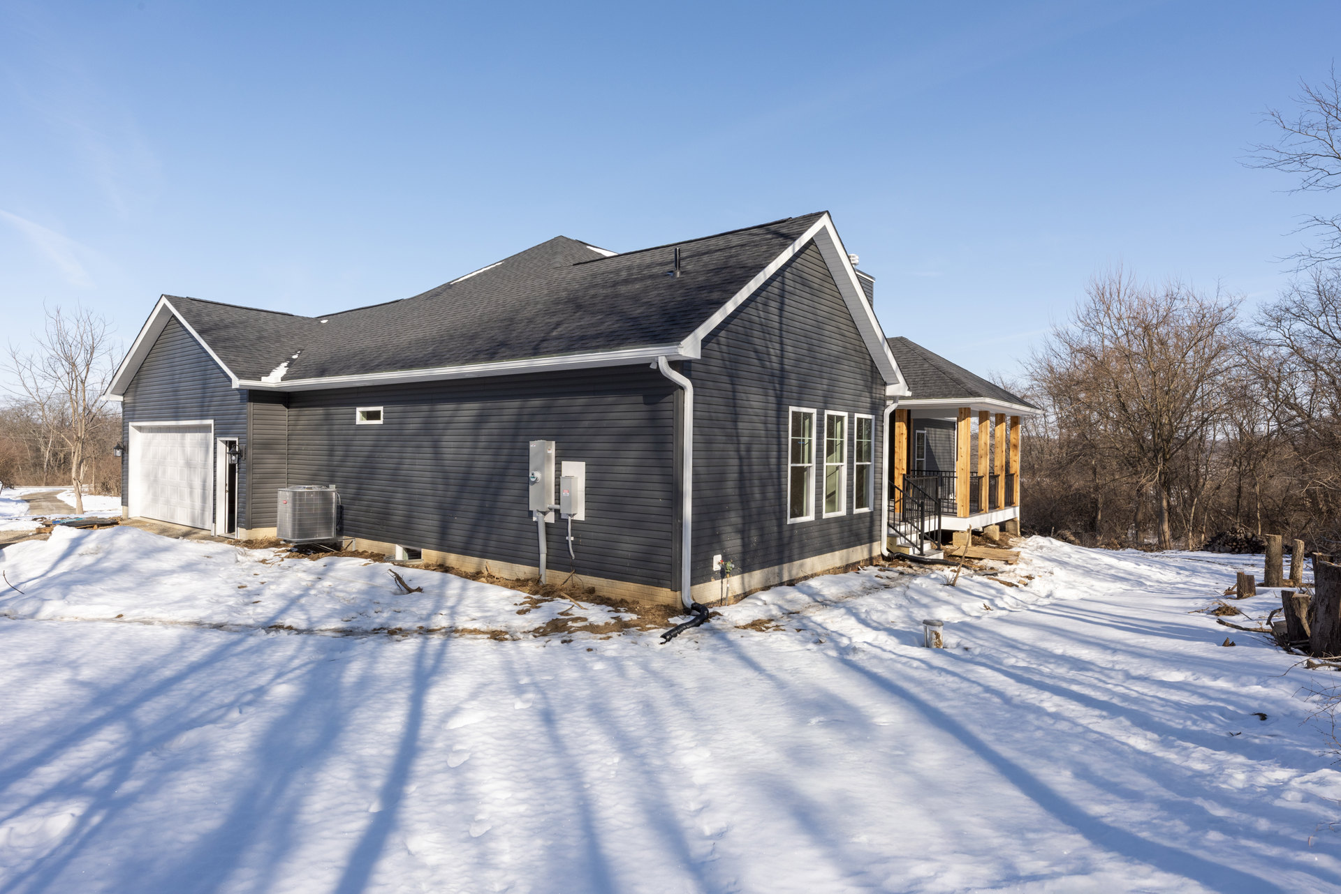 Black-sided house with row of windows, large metal heat pump beside exterior, snow covering ground and roof, black pipe lying near foundation, leafless tree in background under