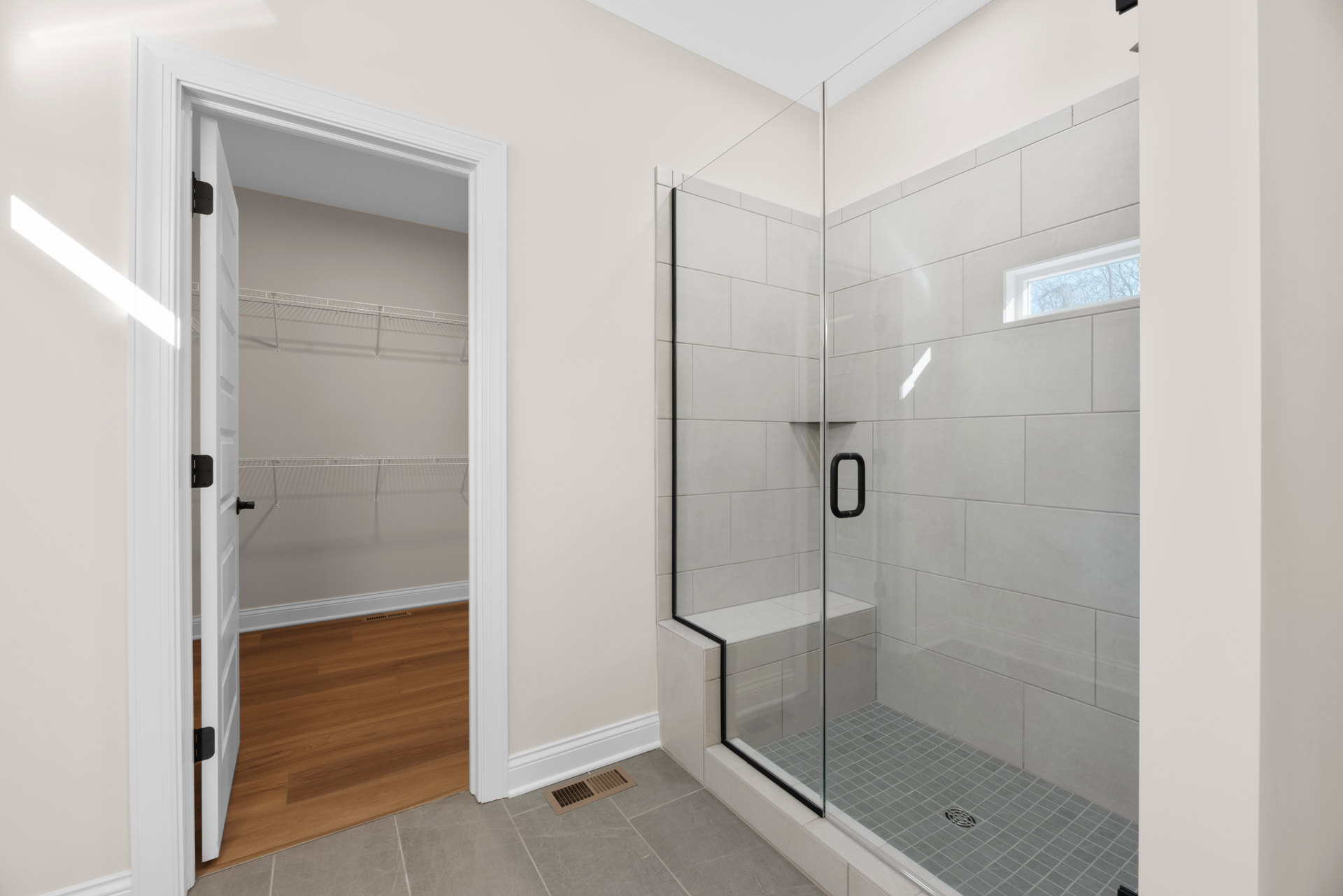 Bathroom with frameless glass shower enclosure, wood plank flooring, open closet door, wall vent, and black metal fixture against white wall