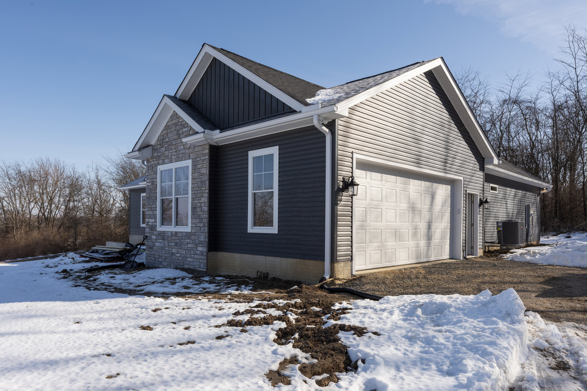 Two-story house with white-framed windows, attached garage, snow covering the ground, bare trees, and a pile of wood and metal near the building