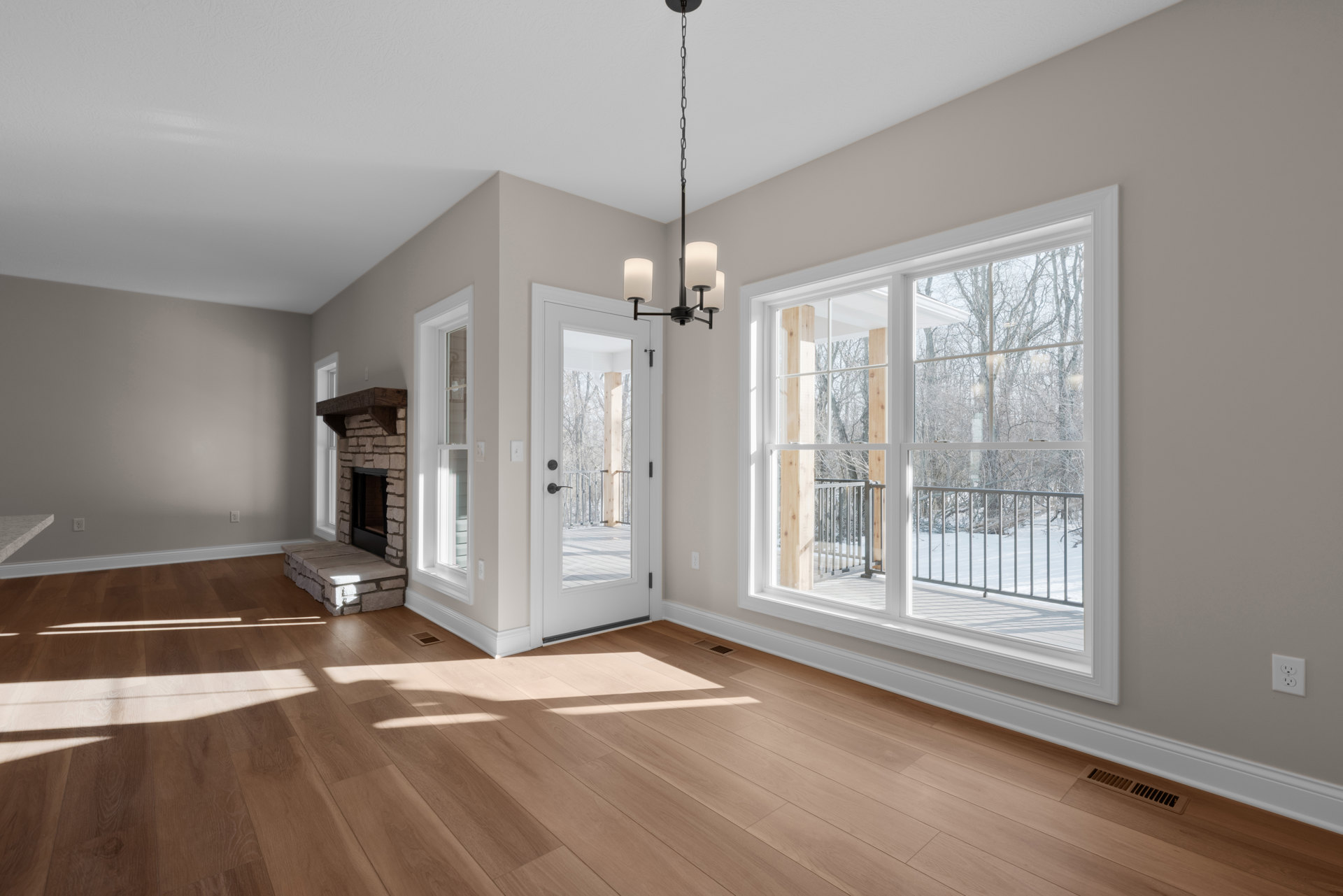 Living room with wood flooring, white walls, fireplace with stone surround, white door opening to deck with trees, large windows with railing, ceiling light fixture hanging from