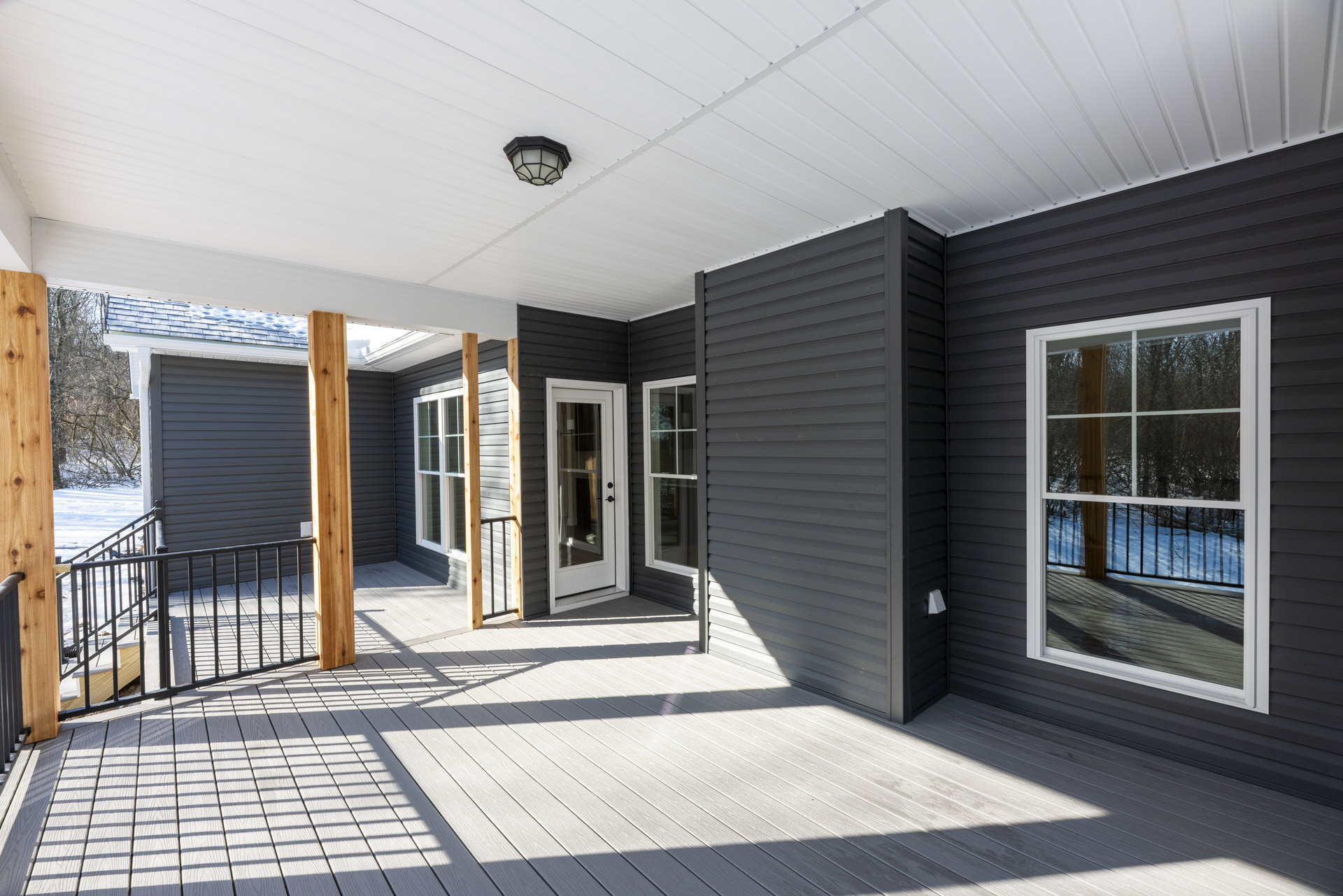 Two-story house with composite deck and white porch railing, glass-paneled white front door, ceiling-mounted light fixture, large window overlooking snowy landscape and trees